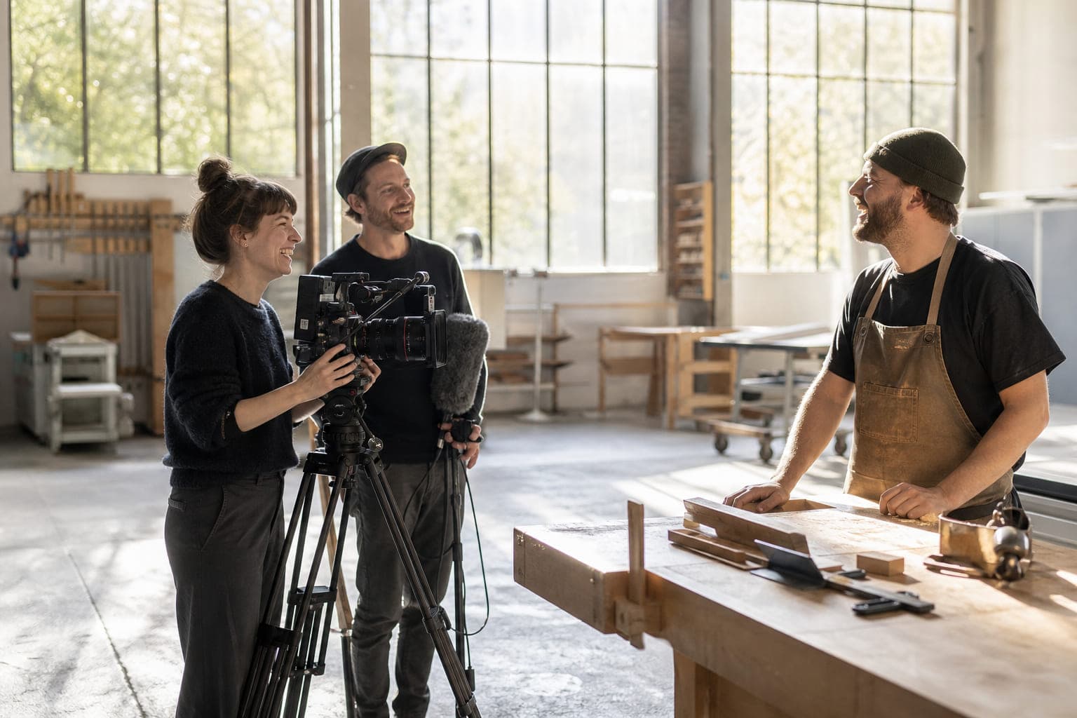 Ein dreiköpfiges Filmteam dreht in einer hellen Werkhalle. Die Kamerafrau lacht mit dem Mitarbeiter, den sie filmt.